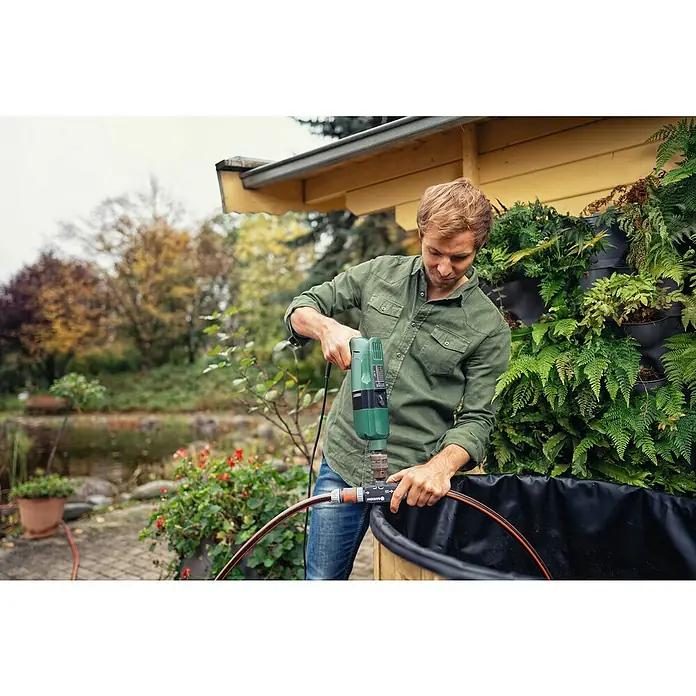 Gardena Pumpe für die Gartenbohrmaschine - Max. Fördermenge: 2.400 l/h, Kunststoff - Mann bohrt mit einem grünen Bohrer ein Loch in ein Bewässerungssystem von Gardena, um es an einen Teich anzuschließen.