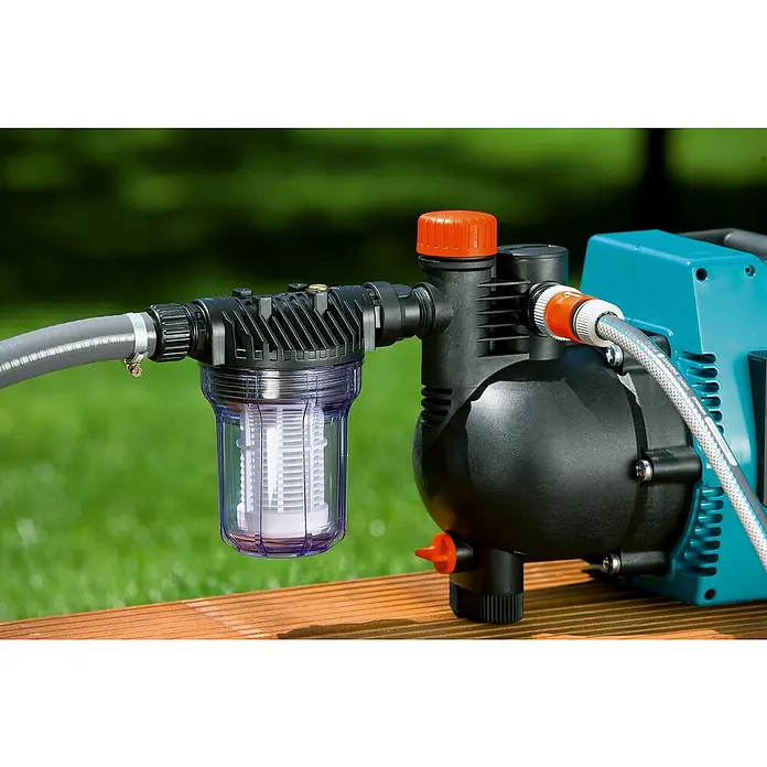 Gardena Hauswasserwerk-Vorfilter mit max. Wasserdurchsatz von 3000 l/h - Größe Anschluss: 33,3 mm (G 1″), Kunststoff - Eine Bewässerungspumpe mit Filter steht auf einem Holzrost im Freien. Der Filter ist transparent und zeigt das Innere.