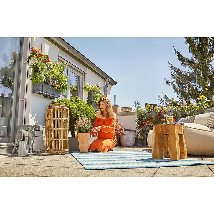 Gardena Wandhalterung NatureUp! - Kunststoff, 2 Stk. - Frau gärtnert auf der Terrasse, umgeben von Pflanzen, die in Töpfen und an Wänden wachsen, während sie ein entspannendes Ambiente genießt.