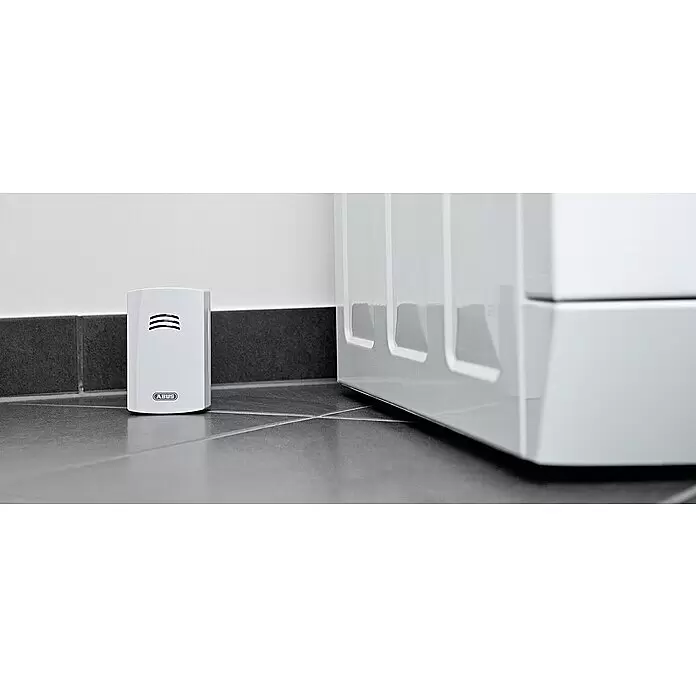 Abus Wassermelder HSWM10000 - Weiß, L x B x H: 35 x 60 x 102 mm - Weißer ABUS Wasserdetektor mit Lüftungsschlitzen auf dunklem Fliesenboden neben einem weißen Gerät.