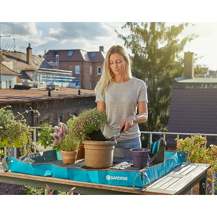 Gardena Pflanzmatte City Gardening - 100 x 120 cm - Eine Frau topft Pflanzen auf einem Tisch mit einer GARDENA Pflanzmatte um. Die Pflanzmatte schützt den Tisch vor Verschmutzung.