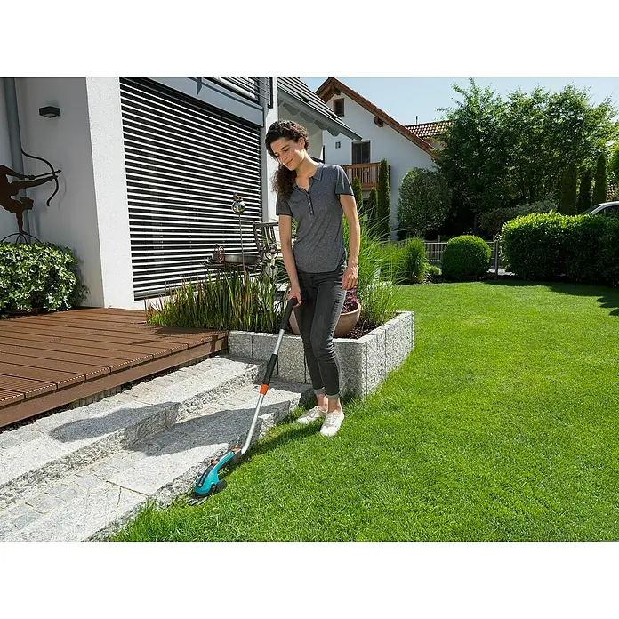 Gardena Teleskop-Komplettroller - Passend für: Gardena Akku-Gras- & Strauchschere - Eine Frau benutzt eine Gardena Akku-Grasschere zur Gartenarbeit. Sie trimmt damit Rasenkanten und Gestrüpp um die Steintreppe herum.