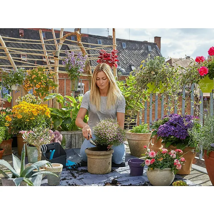 Gardena Pflanzmatte City Gardening - 120 x 180 cm - Frau gärtnert auf Balkon mit Topfpflanzen. Sie pflanzt eine kleine rosa Pflanze in einen Terrakottatopf mit einer Schaufel.