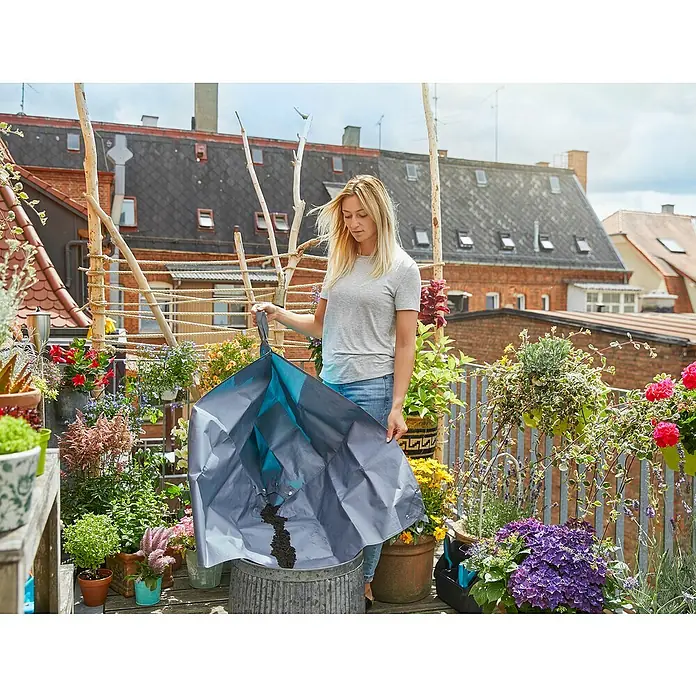 Gardena Pflanzmatte City Gardening - 120 x 180 cm - Eine Frau leert eine Gartentasche in einen Metalleimer, umgeben von üppigen Pflanzen auf einem sonnigen Balkon.