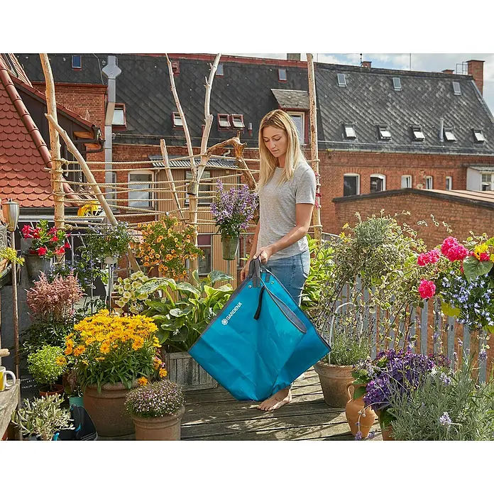 Gardena Formschnittmatte City Gardening - L x B: 150 x 150 cm - Eine Frau steht auf einem Balkon voller Pflanzen und hält eine blaue Gardena Gartentasche.