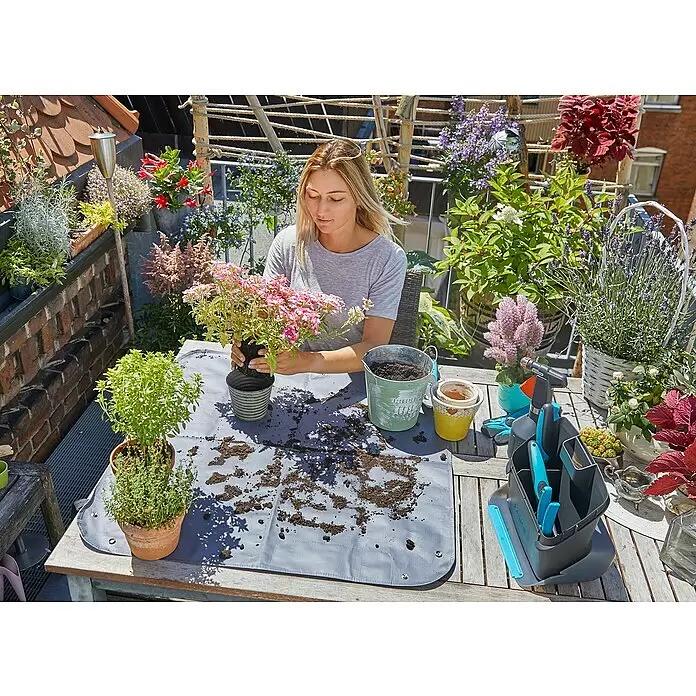 Gardena Pflanzmatte City Gardening - 80 x 80 cm - Eine Frau nutzt eine graue Pflanzmatte auf einem Holztisch zum Umtopfen von Pflanzen, um Erde einzudämmen.