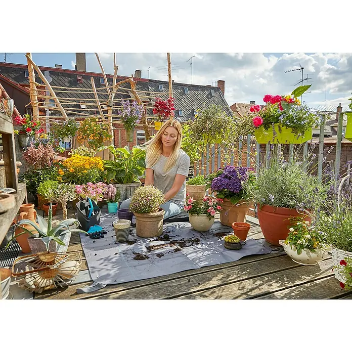 Gardena Pflanzmatte City Gardening - 120 x 180 cm - Eine Frau gärtnert auf einer Dachterrasse, umgeben von einer Vielzahl von Topfpflanzen und Blumen, die eine üppige urbane Gartenoase schaffen.