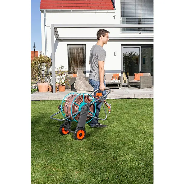 Gardena Schlauchwagen 100 - Metall, Schlauchkapazität: 100 m, 13 mm (1/2") - Ein Mann schiebt einen Gardena-Schlauchwagen mit einem orangefarbenen Schlauch auf einem Rasen vor einem Haus.
