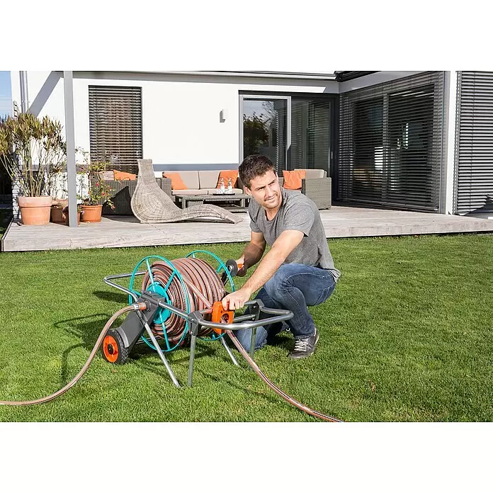 Gardena Schlauchwagen 100 - Metall, Schlauchkapazität: 100 m, 13 mm (1/2") - Mann wickelt einen grau-orangen Gartenschlauch auf eine tragbare Schlauchbox mit blau-grauem Rahmen.