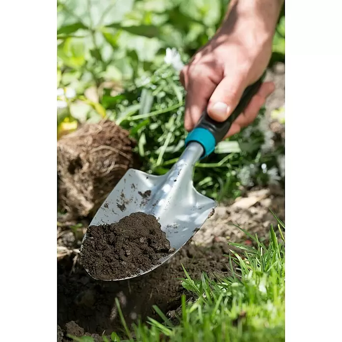 Gardena Comfort Kleingeräte-Set - 3 -tlg. - Eine Hand hält eine silberne Gartenkelle mit schwarzem und türkisfarbenem Griff, gefüllt mit dunkler Erde, beim Graben in einem Garten.