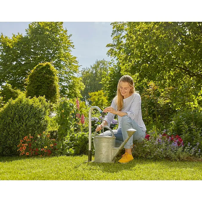Gardena Sprinklersystem Wasserhahn - Metall - Eine Frau befüllt eine Zinkgießkanne mit Wasser im Garten, um Blumen zu gießen, während die Sonne scheint und Bäume im Hintergrund zu sehen sind.