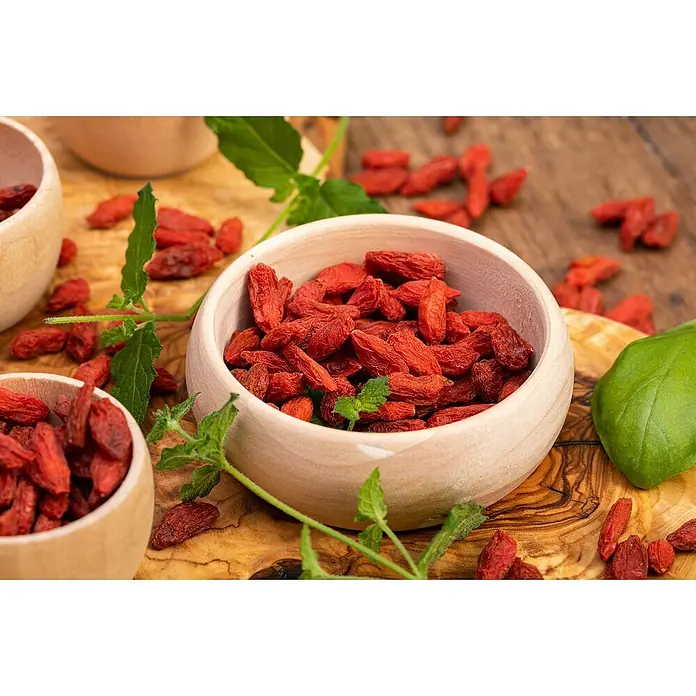Piardino Goji-Beere Bio - Lycium barbarum 'Big and Sweet', Erntezeit: August, Topfvolumen: 2 l - Eine Nahaufnahme von Goji-Beeren in Holzschalen auf einem Holztisch, mit frischen grünen Blättern als dekorative Elemente.