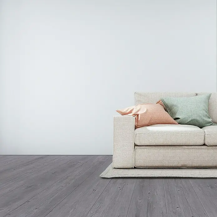 Suelo vinílico SPC Roble Arunda - 1.220 x 180 x 5,5 mm, Gris, Resistente al agua - Primer plano de un sofá beige con cojines verdes y naranjas sobre un suelo de madera gris contra una pared blanca.