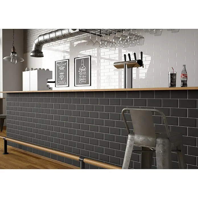 Modern Style Wandfliese Manhattan Biselado Negro - 7,5 x 15 cm, Schwarz, Matt - Die Aufnahme zeigt eine Bar mit einer schwarzen Fliesenfassade, einer hölzernen Theke und verschiedenen Getränken und Gläsern im Hintergrund.
