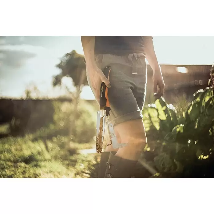Fiskars Xact Hand-Unkrautstecher - 39,6 cm - Person im Garten hält eine Fiskars Xact Gartenschaufel mit orange-schwarzem Griff, von Sonnenlicht beleuchtet.