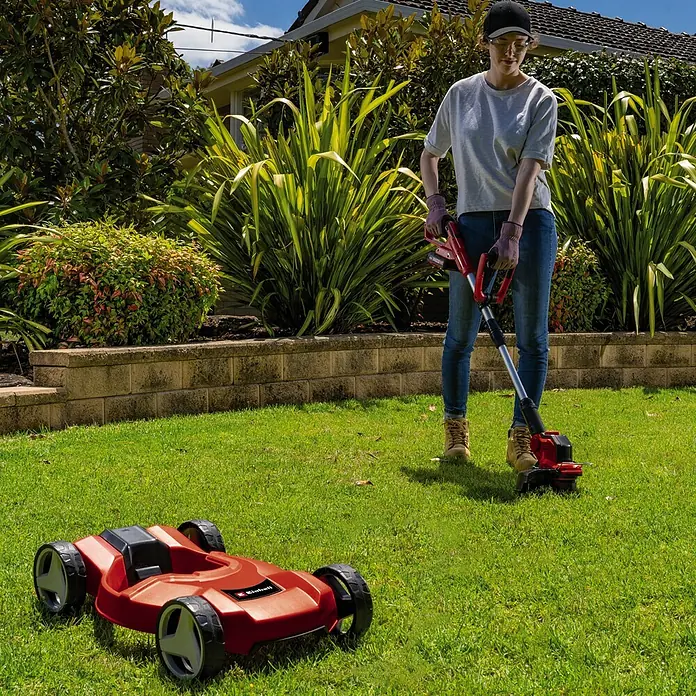 Einhell Power X-Change 18V Akku-Rasentrimmer GE-CT18/28LI TC-Solo18 V, Li-Ionen, Ohne Akku, Schnittbreite Faden: 28 cm - Eine Frau benutzt einen Einhell Akku-Rasentrimmer im Garten, während ein Einhell Mähroboter daneben steht.
