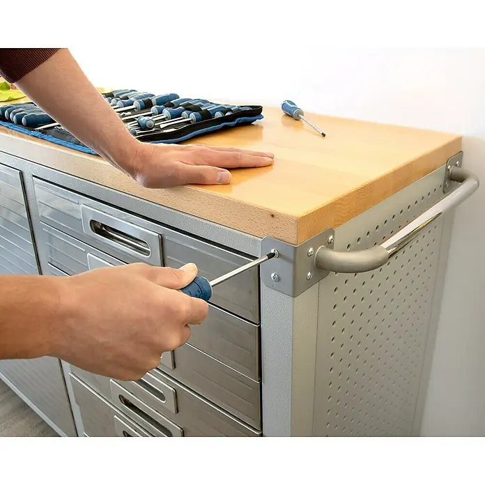 Alpha Tools Schraubendreher-Set - 70 -tlg. - Ein Mann befestigt eine Metallwerkbank mit Holzplatte und Schubladen mit einem Schraubenzieher. Das Werkzeug liegt bereit.