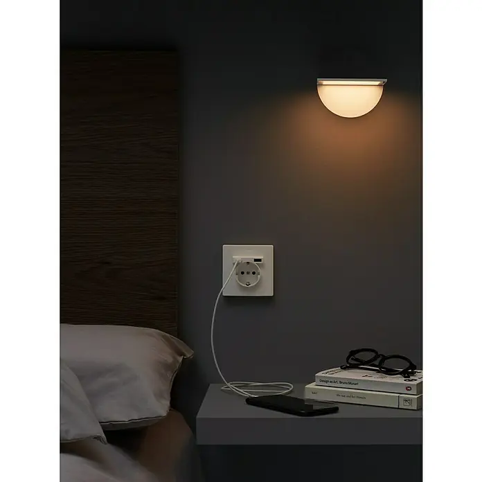 Simon 270 Enchufe con USB - Blanco, 16 A - Una lámpara de pared está montada sobre una mesita de noche con un libro y gafas, proyectando una luz cálida. El ambiente es tranquilo y moderno.