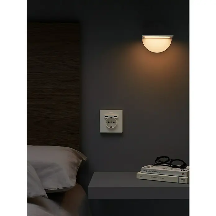 Simon 270 Enchufe con USB - Blanco, 16 A - Lámpara de pared sobre la mesita de noche con toma USB y dos libros. La iluminación suave crea un ambiente acogedor en el dormitorio.