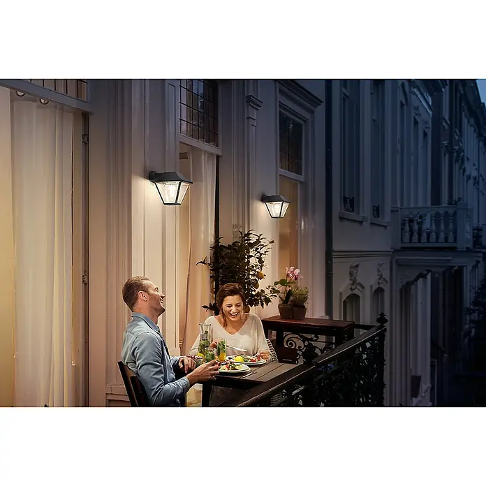 Philips Alpenglow Aplique exterior - 60 W, 17,4 x 30 x 17,9 cm, Antracita, IP44 - Una pareja disfruta de una cena en el balcón, iluminado por elegantes luces de exterior montadas en la pared, creando un ambiente acogedor.