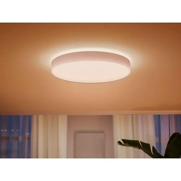 Philips Hue LED-Deckenleuchte rund White Ambience Enrave XL - 48 W, Ø x H: 55,1 x 8,4 cm, Weiß, Kaltweiß - Nahaufnahme einer runden LED-Deckenleuchte, die warmes Licht in einem Raum mit Vorhängen und Zimmerpflanzen spendet.