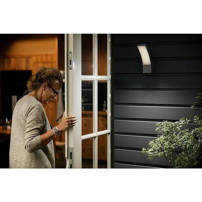 Philips Splay Aplique exterior LED - 12 W, L x An x Al: 16 x 8 x 24 cm, Color del marco: Antracita, Blanco frío - Una mujer abre una puerta que presenta una luz exterior elegante, enfatizando el diseño moderno de la casa.