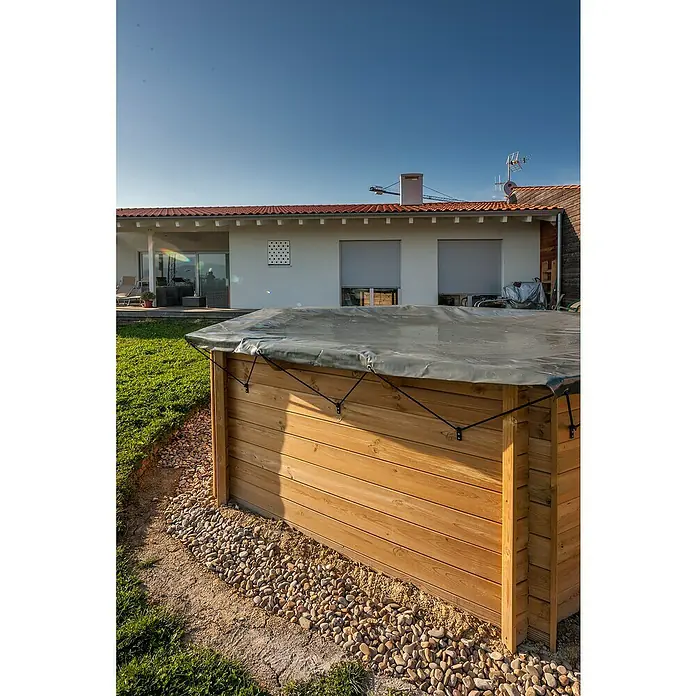 Gre Pool-Abdeckplane Winter Vanille - 580 g/m², Passend für: Pools bis Ø 400 cm - Außenaufnahme einer hölzernen Badezuber-Umrandung mit einer grauen Abdeckung, umgeben von Steinen, vor einem Haus an einem sonnigen Tag.