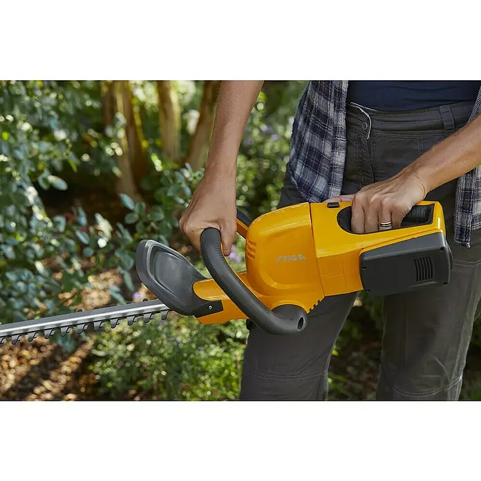 Stiga 100er Serie Akku-Heckenschere HT 100e Set - 20 V, Li-Ionen, 1 Akku, Schnittlänge: 50 cm - Nahaufnahme einer gelben STIGA-Heckenschere, die von einer Person in Arbeitskleidung gehalten wird. Ideal zum Trimmen von Hecken im Garten.