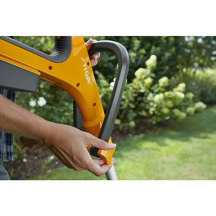 Stiga 100er Serie Akku-Rasentrimmer GT 100e - Set - 20 V, 2 Ah, Li-Ionen, 1 Akku, Schnittbreite Faden: 25 cm - Eine Person hält einen gelben STIGA-Rasentrimmer in der Hand, der sich vor einem grünen Garten befindet.