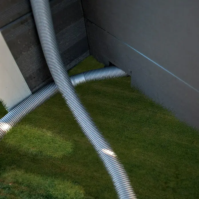 Gre Technikraum - L x B x H: 80 x 60 x 115 cm - Nahaufnahme von flexiblen Lüftungsschläuchen aus Aluminium, die auf Rasen vor einer Gebäudeaußenwand liegen.
