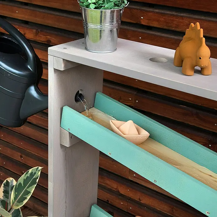 Muddy Buddy Wasserbahn Auqa Tower - Holz, Salbei - Nahaufnahme einer Outdoor-Wasserspielstation mit Gießkanne, Eimer, Spielzeugdino und einem Papierboot, das einen Kanal hinuntertreibt.