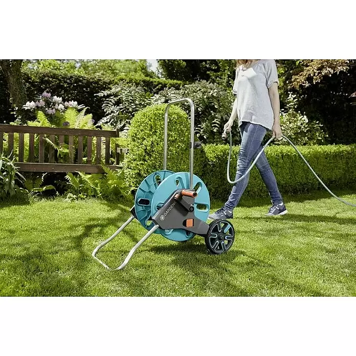 Gardena Schlauchwagen CleverRoll M - Kunststoff/Metall, Schlauchkapazität: 60 m, 13 mm (½″) - Eine Person zieht eine blau-graue Gardena Schlauchbox auf Rädern über eine grüne Wiese und hält den Schlauch.