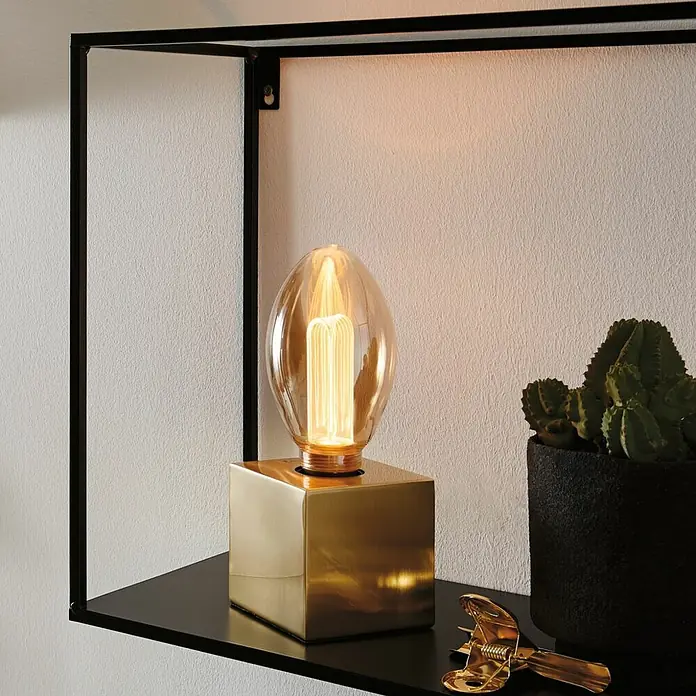 Paulmann Inner Glow LED-Lampe Vintage Glühlampenform E27 - E27, Nicht Dimmbar, Gelb, 160 lm, 3,5 W, Kerze, Farbe: Gold - Nahaufnahme einer Tischlampe mit einem goldenen Sockel und einer dekorativen Glühbirne auf einem Regal, neben einer Kaktuspflanze.