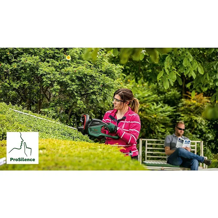 Bosch Elektro-Heckenschere UniversalHedgeCut 60 - 480 W, Schnittlänge: 60 cm - Eine Frau schneidet mit einer Bosch Heckenschere eine Hecke, während ein Mann im Hintergrund liest. Die Heckenschere ist mit ProSilence Technologie ausgestattet.