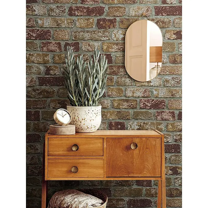 Marburg Sand & Stones Vliestapete Steinmauer II - Beige, Steinoptik, 10,05 x 0,53 m - Dekorative Nahaufnahme eines Holzschranks mit Pflanze, Uhr und Spiegel vor einer Ziegelwand.