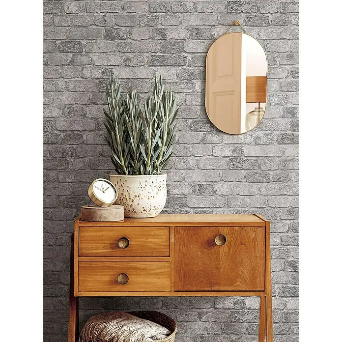 Marburg Sand & Stones Vliestapete Steinmauer II - Grauweiß, Steinoptik, 10,05 x 0,53 m - Stilvolles Interieur mit einem Holzschrank, einer Pflanze und einem Spiegel vor einer grauen Backsteinwand.