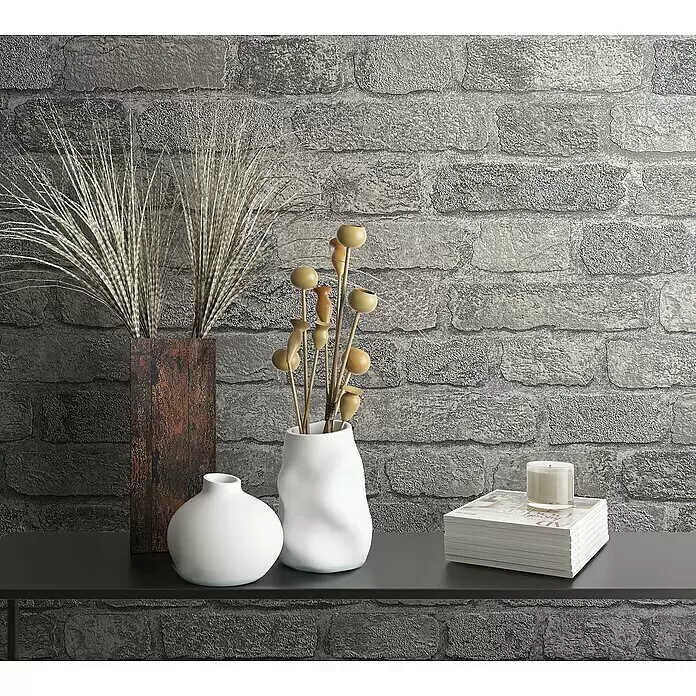 Marburg Sand & Stones Vliestapete Steinmauer II - Grauweiß, Steinoptik, 10,05 x 0,53 m - Ein dunkles Regal mit Deko-Artikeln: rostige Vase mit Trockengras, zwei weiße Vasen mit Kapseln, Kerze auf Zeitschriften, vor Ziegelwand.