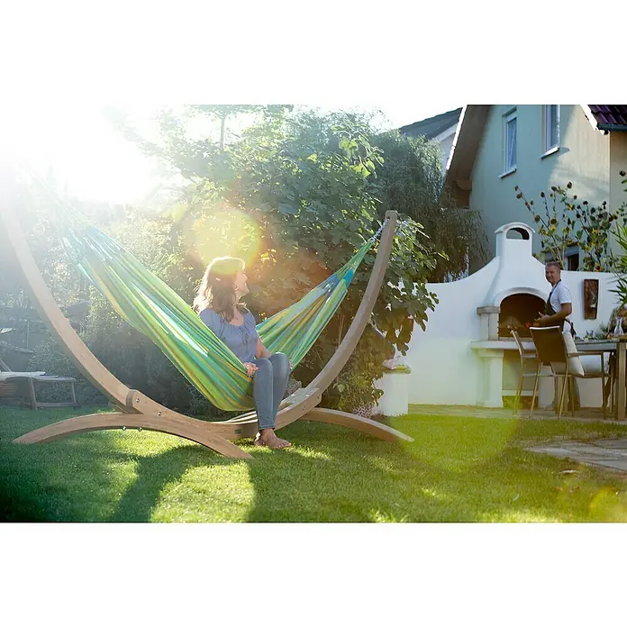 La Siesta Hängematte Brisa - L x B: 350 x 160 cm, Lime - Frau entspannt in einer Hängematte auf einem Holzgestell im Garten, während im Hintergrund gegrillt wird.