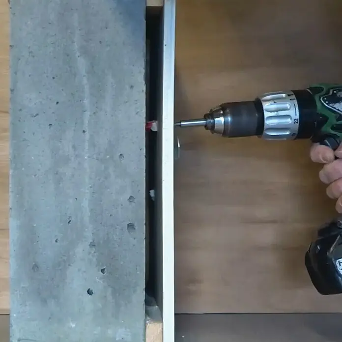 Corefix Trockenbau-Set 100 - Länge Schraube: 100 mm - Ein grüner Akku-Bohrer wird verwendet, um eine Metallhalterung an einer Betonwand zu befestigen, die von einem Holzrahmen umgeben ist.