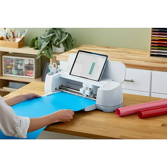 Cricut Maker Schneideplotter Maker 3 - Blau, Bluetooth, Schnittbreite: 29,7 cm, Schnittlänge: 3,6 m - Nahaufnahme eines Cricut Maker 3 Schneideplotters mit einer Person, die blaues Material zuführt, und einem Tablet, das ein Design anzeigt.