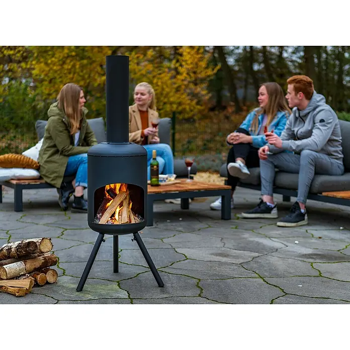 RedFire Gartenkamin Fuego - Ø x H: 40 x 148 cm, Stahl - Ein schicker, schwarzer Außenkamin steht auf einer Terrasse, während vier Personen in der Nähe sitzen und sich unterhalten.