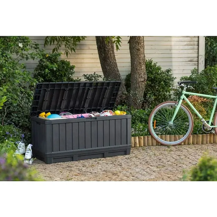 Keter Arcón para cojines de exterior Kentwood - L x An x Al: 128 x 53,6 x 59 cm, Plástico, Gris - Una caja de almacenamiento exterior gris con tapa abierta, llena de varios artículos, está en el jardín al lado de una bicicleta verde.