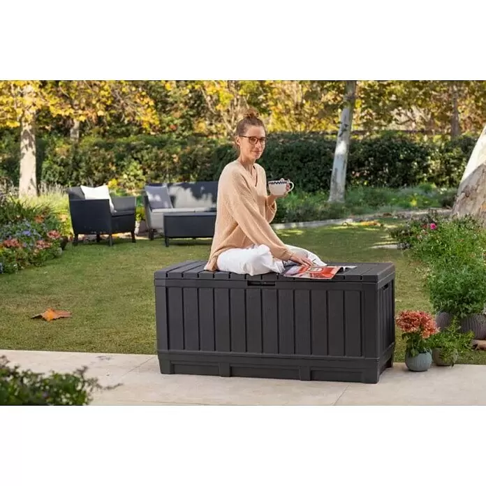 Keter Garten-Aufbewahrungsbox Kentwood - L x B x H: 128 x 53,6 x 59 cm, Kunststoff - Dunkelgraue Outdoor-Aufbewahrungsbox als Gartenbank genutzt, mit einer darauf sitzenden lesenden Frau.