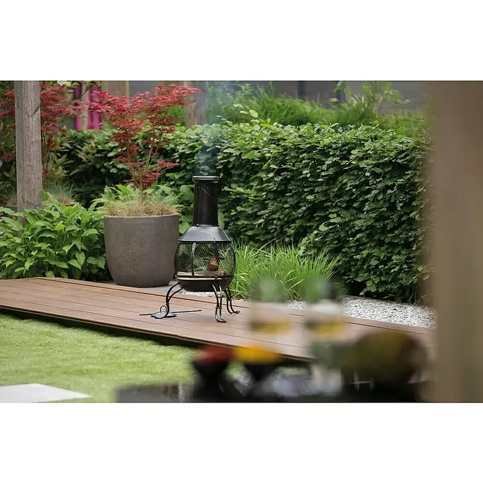 RedFire Gartenkamin Sauda - Ø x H: 38 x 90,5 cm, Stahl - Gemütliche Gartenszene mit einem schwarzen Kaminofen, der auf einer Holzterrasse steht, umgeben von üppigem Grün.