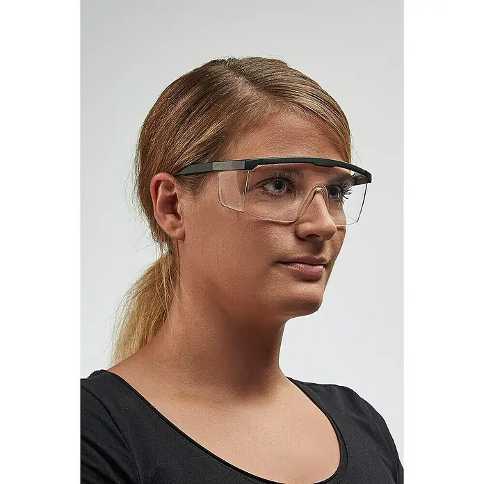 Schutzbrille - Verstellbare Bügel, Blau - Eine Frau trägt eine transparente Schutzbrille mit schwarzem Rahmen, die ihre Augen vor Staub und Spritzern schützt. Ideal für Werkstatt und Labor.
