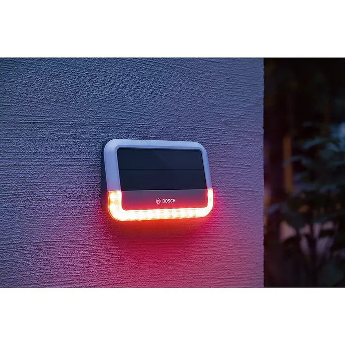 Bosch Smart Home Außensirene - 100 dB - Nahaufnahme einer Bosch Außenleuchte mit Solarpanel, die an einer strukturierten Wand montiert ist und rotes Licht ausstrahlt.