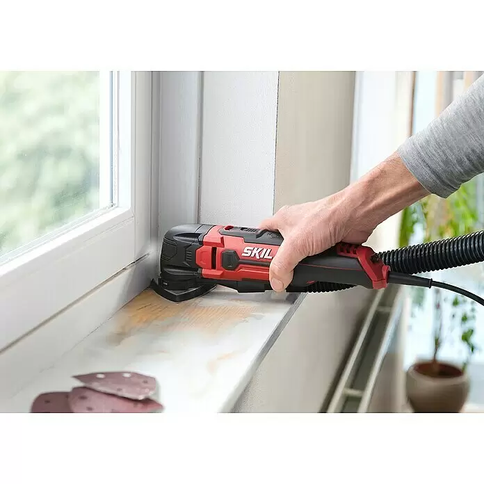 Skil Elektro-Multifunktionswerkzeug 1491 DB - 300 W - Rotes und schwarzes SKIL Multifunktionswerkzeug schleift eine hölzerne Fensterbank, von einer Hand gehalten.