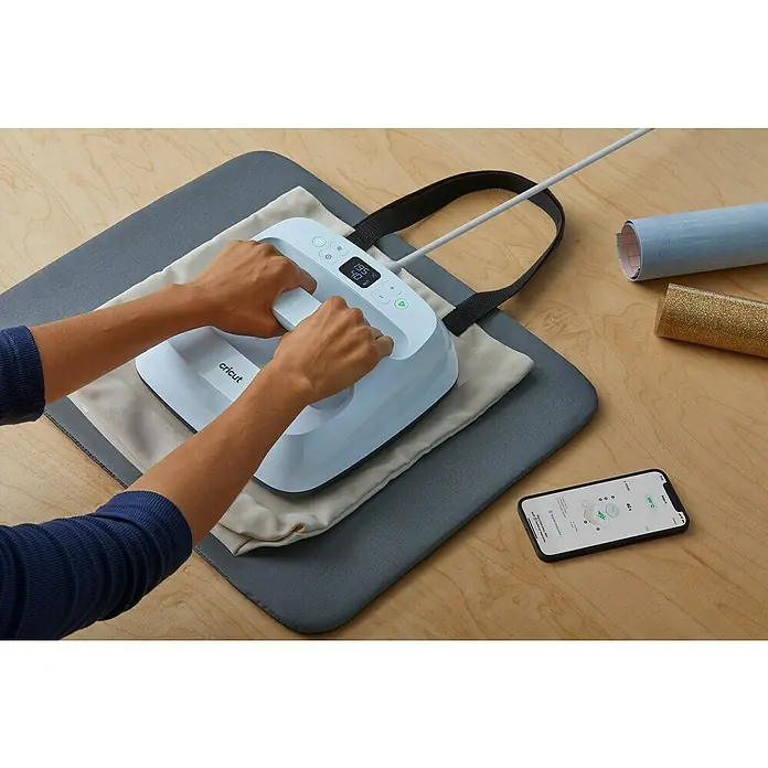 Cricut Transferpresse Easypress 3 - B x L: 23 x 23 cm, Passend für: Bügelfolien, Drucktechnologie: Wärmeübertragung - Cricut EasyPress 3 wird auf einer grauen Bügelmatte mit einer Tragetasche und einer Rolle Vinyl verwendet. Ein Smartphone liegt daneben.