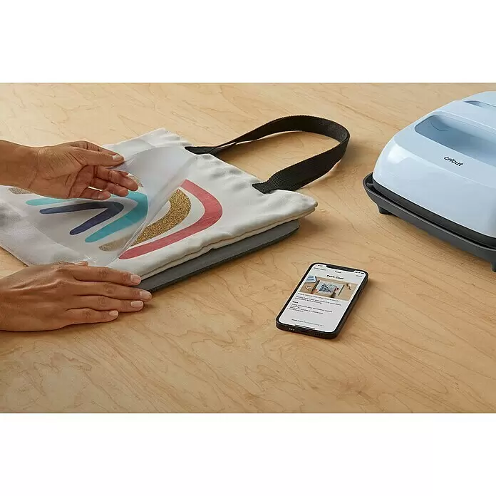 Cricut Transferpresse Easypress 3 - B x L: 23 x 23 cm, Passend für: Bügelfolien, Drucktechnologie: Wärmeübertragung - Hände lösen Transfer von Tragetasche mit Design, geleitet vom Smartphone. Eine Cricut-Heißpresse ist auf Holzfläche sichtbar.