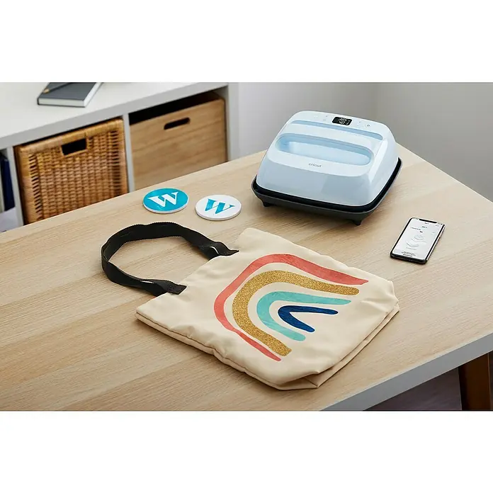 Cricut Transferpresse Easypress 3 - B x L: 23 x 23 cm, Passend für: Bügelfolien, Drucktechnologie: Wärmeübertragung - Kreative Arbeitsfläche mit Cricut EasyPress 3, Smartphone und einer Tasche mit Regenbogenmotiv für DIY-Projekte.